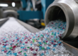 Close-up of colorful, high-purity recycled plastic pellets pouring from a metallic chute onto a conveyor belt in a validated decontamination facility.