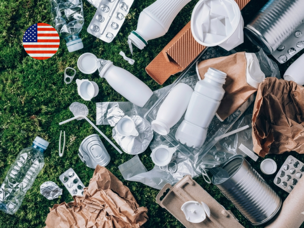 Assorted plastic, paper, and metal packaging waste on a green grass background with a U.S. flag icon, illustrating the 2026 U.S. EPR reporting requirements and SB 54 compliance for producers.