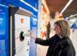 Consumer returning a reusable cup to a TOMRA automated return machine inside a busy German train station.