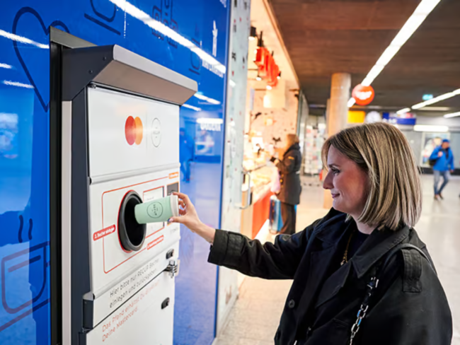 Consumer returning a reusable cup to a TOMRA automated return machine inside a busy German train station.