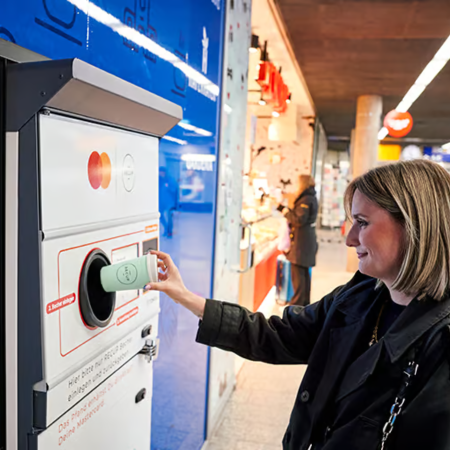 Consumer returning a reusable cup to a TOMRA automated return machine inside a busy German train station.