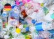 Mixed plastic packaging waste including bottles, containers and flexible materials with a German flag icon, illustrating the proposed German plastics tax (Plastikabgabe) and industry opposition to the budget-driven measure.