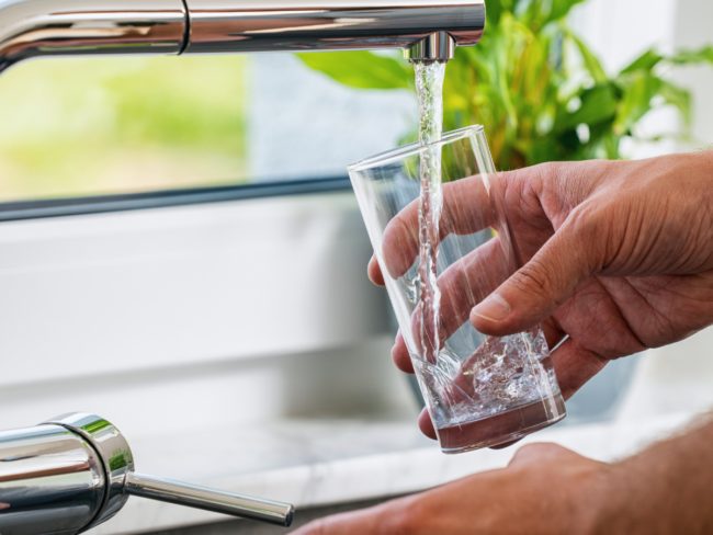 Glass being filled with drinking water from a kitchen tap, illustrating the new EU rules for products in contact with drinking water from 31 December 2026.